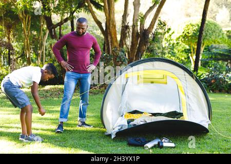 Il padre africano americano che guarda il figlio pitching tenda in giardino soleggiato Foto Stock