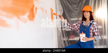 Felice donna caucasica in elmetto dipinge il muro interno in una nuova casa con un pennello in arancione. Concetto di costruzione e ristrutturazione. Ristrutturazione della casa Foto Stock