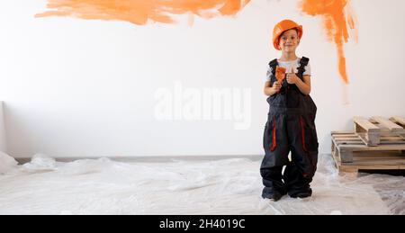 Riparazione chiavi in mano. Ragazzino in casco e tute con un pennello sullo sfondo di una parete bianca con vernice arancione. Nuova ristrutturazione della casa Foto Stock