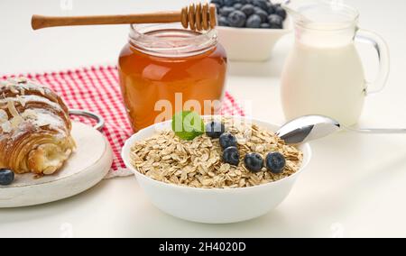 Colazione al mattino, fiocchi d'avena crudi in un piatto di ceramica, latte in un decanter, mirtilli e miele in un vaso su un tavolo bianco Foto Stock