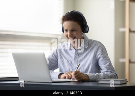 Sorridente donna matura in cuffie che studia su corsi online. Foto Stock