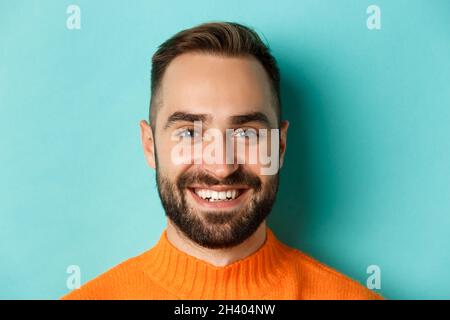 Headshot di bell'uomo caucasico con barba sorridente felice alla macchina fotografica, in piedi in pullover arancione contro sfondo turchese Foto Stock