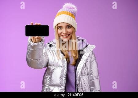 Ragazza bionda sicura e amichevole all'aperto con giacca glittering argento cappello tenere smartphone orizzontale mostrando pho mobile Foto Stock