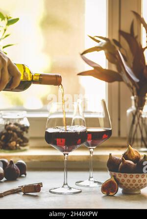 Versare a mano il vino rosso dalla bottiglia in due bicchieri da vino rosso sul tavolo da cucina con fichi, posate di vino e sughero sullo sfondo della finestra. Luce naturale. Dal Foto Stock