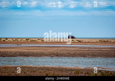 Un colpo lontano di un trattore rosso sulla spiaggia a Gibilterra Point, Lincolnshire, Regno Unito con ciuffi di canne e un fiume che scorre in primo piano. Foto Stock