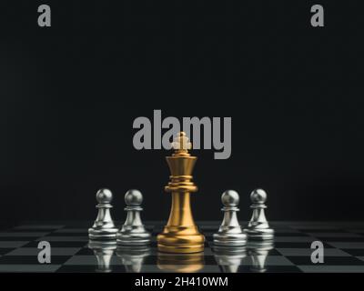 Pezzi di scacchi del re d'oro in piedi al centro di piccoli pezzi di pedine d'argento su scacchiera su sfondo scuro. Giochi di scacchi figura in gioco di scacchi com Foto Stock