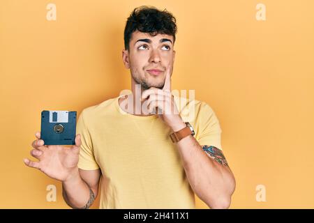 Giovane ispanico che tiene il floppy disk serio faccia pensando alla domanda con mano sul mento, pensata per un'idea confusa Foto Stock