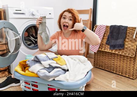 Giovane donna rossa che mette la lavanderia sporca in lavatrice sorridendo facendo il gesto del telefono con la mano e le dita come parlare al telefono. commun Foto Stock