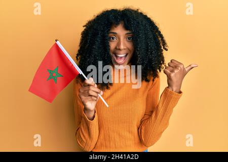 Donna afroamericana con capelli afroamericani che tengono bandiera del marocco puntando il pollice sul lato sorridendo felice con bocca aperta Foto Stock
