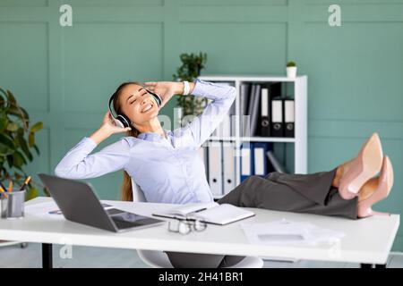 La vita è buona. Gioiosa imprenditrice femminile che ascolta musica sul posto di lavoro e mette i piedi sul tavolo Foto Stock