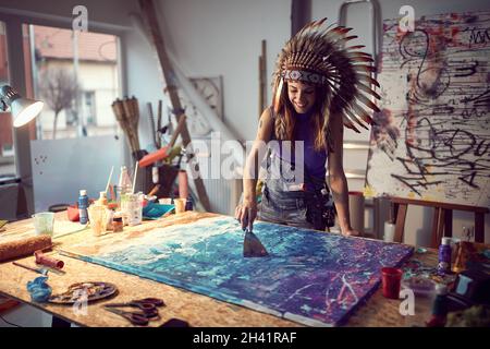 Una giovane artista femminile con un cofano di guerra sta usando una spatola per spargere la vernice sopra il suo nuovo dipinto che sta lavorando in un'atmosfera piacevole nel Foto Stock