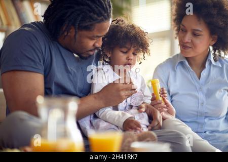 Una ragazza piccola carina godendo di passare il tempo con i suoi genitori e fare le bolle di sapone in un'atmosfera giocosa a casa. Famiglia, insieme, tempo di gioco Foto Stock