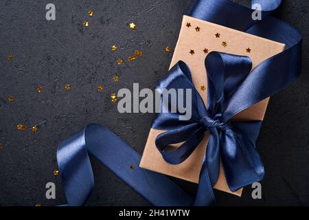 Confezione regalo per le feste o regalo con nastro blu, confetti dorati e baubles d'oro su sfondo nero. Biglietto d'auguri magico di natale. Decorazione di Natale Foto Stock