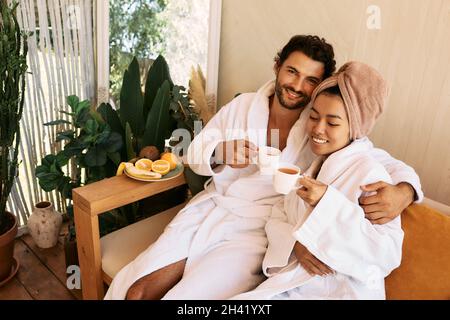 Una giovane coppia amorevole in accappatoi bianchi passa il tempo al centro benessere e gusta un tè benessere. Coppia durante un week-end romantico Foto Stock