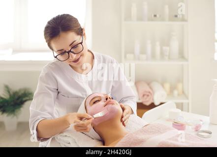 Estetista al salone di bellezza che applica maschera facciale con argilla rosa sul viso della giovane donna Foto Stock