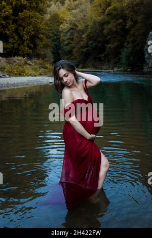 Giovane, elegante, donna incinta bionda in piedi nel fiume in abito rosso Foto Stock