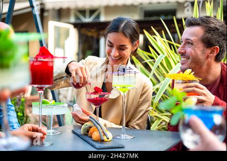 Amici del gruppo che si divertono bevendo bevande colorate seduti nel cortile del bar - ragazza felice sorridendo e versa la pozione rossa nel bicchiere da cocktail - bere Foto Stock