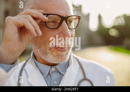 Primo piano ritratto di fidato medico di sesso maschile in camice da laboratorio di regolazione dei suoi occhiali, guardando via, posa all'aperto vicino alla clinica Foto Stock