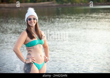 Una giovane donna che indossa un bikini in piedi vicino al bordo del mare e indossa un cappello di lana pronto per una nuotata degli inverni Foto Stock