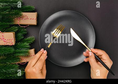 Vista dall'alto delle mani donna che tengono coltello e forchetta su piatto nero vuoto con telaio di rami di albero di Natale Foto Stock