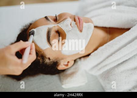 Il beautician applica la maschera di faccia di argilla bianca con un pennello sul volto di una donna. Foto Stock