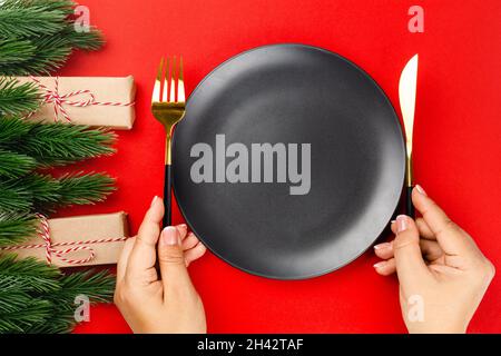 Donna mani tenere coltello e forchetta vicino nero piatto vuoto su tavola rossa con alberi di Natale e decorazioni Foto Stock