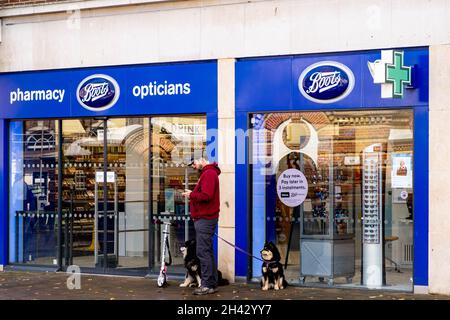 Epsom Surrey London UK, ottobre 31 2021, Boots Pharmacy and Opticians Retail Store Shop Front con logo con uomo tramite telefono cellulare e due cani da compagnia S Foto Stock