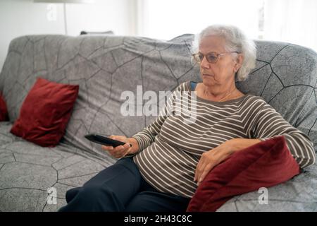 Donna matura che tiene il telecomando della TV mentre guarda la televisione a casa Foto Stock