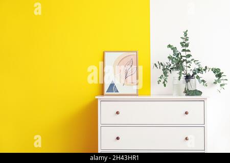 Vaso con rami di eucalipto e quadro su cassettiera vicino a muro colorato Foto Stock