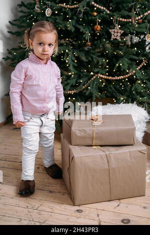 Carino bambino toddler in attesa di regali a casa dopo la decorazione albero di Natale in stile ecologico con giocattoli di legno. Serata natalizia per bambini. Stile nordico Foto Stock