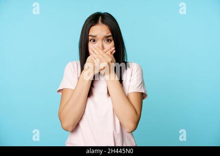 Scioccata donna asiatica copre bocca urla, gaspando affettato, spaventato di smth, chiude le sue labbra, in piedi su sfondo blu. Foto Stock