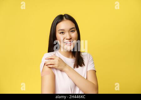 Covid-19 e il concetto medico sanitario. Donna sorridente mostra spalla con cerotto, vaccinato da coronavirus, in piedi in t-shirt sopra il giallo Foto Stock