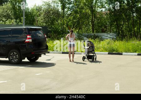 Una ragazza con un passeggino nel parcheggio. Una donna si riposa vicino all'auto. Una persona dopo un lungo viaggio si trova sulla strada. Passeggino per camminare con un ch Foto Stock