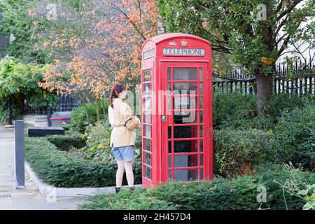 Una donna si erge fuori da un telefono rosso sulla strada di Londra circondata da colori autunnali di foglie di albero e piante verdi giardino Inghilterra UK Foto Stock