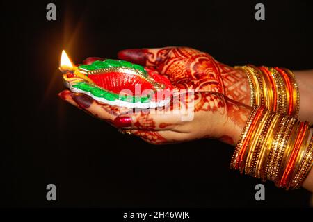 Donna mano che indossa mehndi e sanghette in primo piano tenendo una lampada Diwali Diya su sfondo scuro Foto Stock