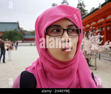 Primo piano ritratto della giovane donna musulmana asiatica che indossa hijab e occhiali nel cortile del santuario in Giappone. Eccitato, felice e anatra espressione del volto. Foto Stock