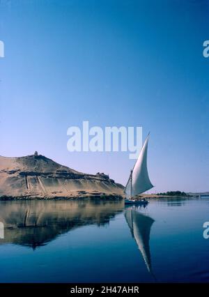 Africa, Egitto, Luxor, fiume Nilo 1976. Una feluca sul Nilo ad Assuan. Tomba sulla collina di Qubbet el Hawa e antiche tombe nella scogliera del deserto. Foto Stock