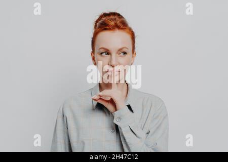 Giovane donna zenzero che tiene il dito sulle labbra e guarda da parte mentre si alza contro il muro grigio Foto Stock