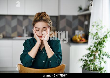 Ritratto di giovane studentessa stanca seduta e rilassante con gli occhi chiusi in cucina. Foto Stock