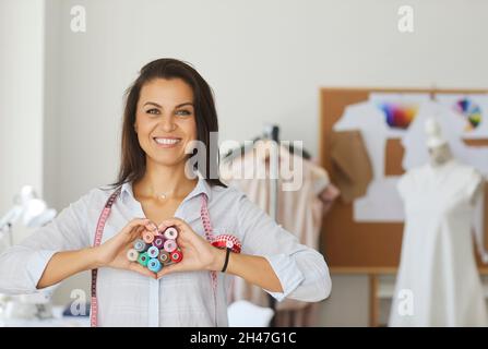 Ritratto di donna amichevole seamstress mostrando le mani cuore a macchina fotografica in laboratorio di cucito. Foto Stock