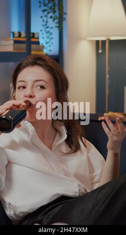 Persona che mangia una fetta di pizza mentre guardi la commedia in TV e ride sul divano a casa. Donna caucasica divertirsi con film in televisione, gustando fast food takeaway e bottiglia di birra Foto Stock
