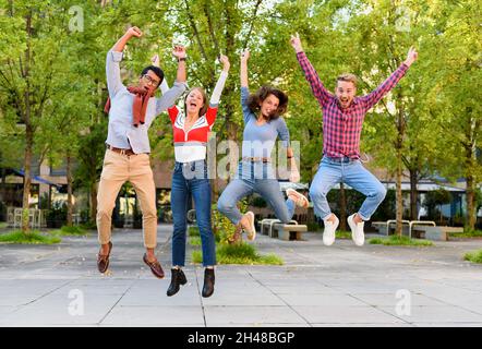 Gruppo di giovani amici esuberanti e diversi che allietano e saltano in aria con le braccia sollevate in festa all'aperto in un parco Foto Stock