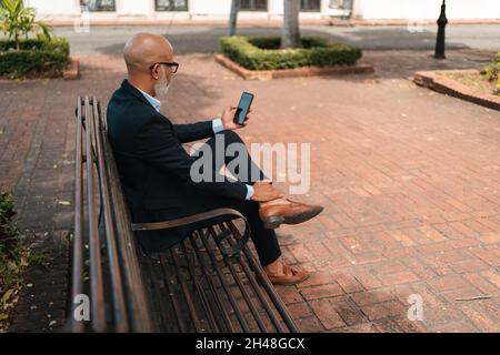 Bald maschio ispanico in un abito elegante utilizzando il suo smartphone mentre si siede su una panca Foto Stock