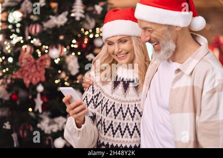 Coppia matura felice in cappelli di santa usando il telefono delle cellule vicino all'albero di Natale nel paese. Foto Stock