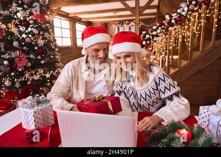 Felice vecchia coppia indossare babbo natale cappelli tenendo regalo saluto famiglia su festa virtuale. Foto Stock