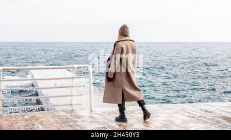 Donna elegante che indossa un lungo cappotto beige, stivali neri chunky e cappello in maglia in piedi da solo sul molo guarda il mare in una fredda giornata invernale. Foto Stock