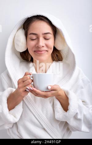 Bella donna piacevole tenendo la tazza in mani, bere tè o caffè, isolato su sfondo bianco, indossare accappatoio godendo con gli occhi chiusi, sorridente.Sp Foto Stock