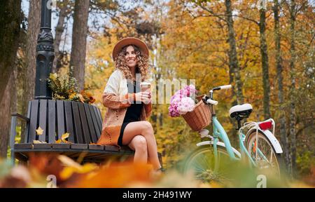 Vista frontale della bella donna con bicicletta retrò e tazza di caffè è seduta su panca di legno. La femmina curly sta riposando sulla sistemazione nel parco d'autunno. Concetto di fine settimana. Foto Stock