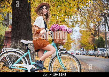 Giro in bicicletta in autunno attraverso la città. Bella donna sorridente ciclista con capelli ricci in elegante cappello in bicicletta retrò che si ferma vicino all'albero su una delle strade della città durante la bicicletta nel fine settimana. Foto Stock