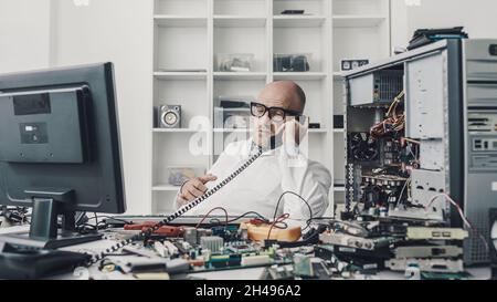 Un tecnico distratto seduto alla scrivania e parlando al telefono invece di riparare un computer rotto Foto Stock